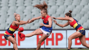 Three AFLW players, the player in the middle is attempting to kick away the ball