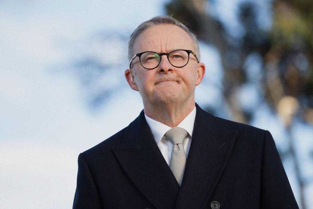 Prime Minister Anthony Albanese looks onwards facing the camera, dressed in a black coat, white shirt and patterned grey tie. He is wearing glasses and is grimacing in this photo.