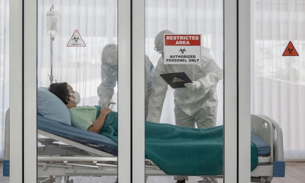 A woman lies in a hospital bed, surrounded by two medical workers dressed in white protective medical PPE. All subjects are in an enclosed room. A sign on the front window reads, 'Restricted Area: Authorised Personnel Only'.

