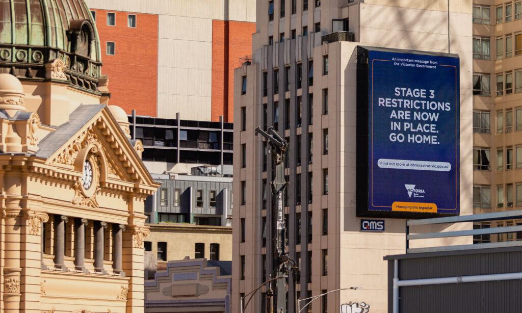 A photo of Flinders Street, Melbourne during the Victorian COVID-19 lockdown. In focus, the Flinders Street Station facade, and a digital billboard that reads, 'Stage 3 Restrictions are now in place. Go home'. 