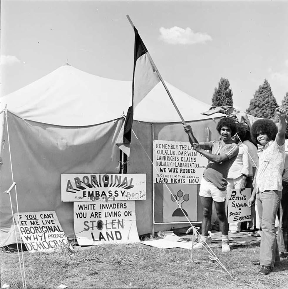 This is a black-and-white photograph of activists at the Aboriginal Tent Embassy on the lawns of (Old) Parliament House in Canberra in 1974. The photograph shows placards placed against a canvas tent and a few men standing in front of the tent. One man is waving an Aboriginal flag