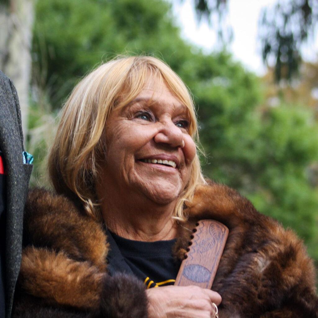 Bangerang and Wiradjuri Elder and Co-Chair of the First People's Assembly of Victoria, Aunty Geraldine Atkinson looks out to the camera. She is wearing a shawl made of fur, smiling and holding a wooden object.