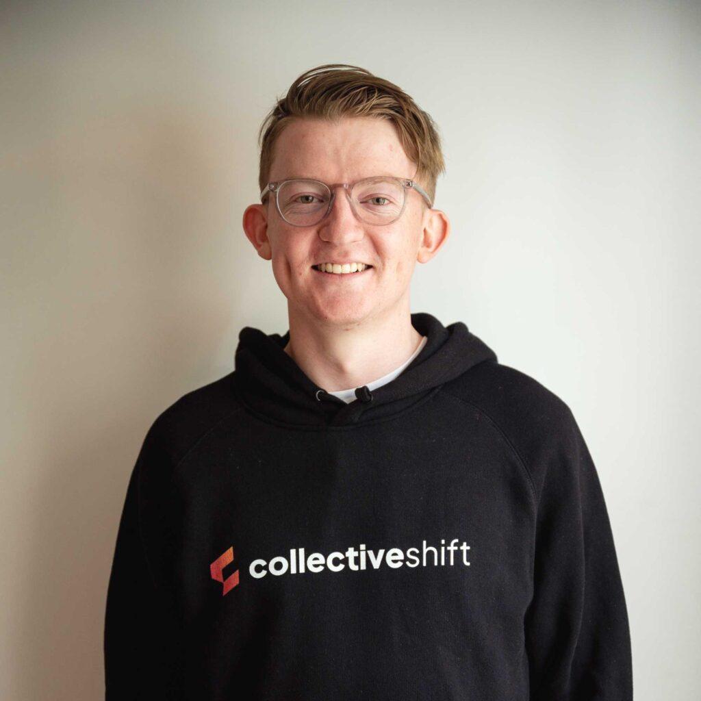 Collective Shift founder Ben Simpson stands facing the camera against a beige background. He is wearing glasses, smiling and wearing a black hoodie with the words 'Collective Shift' across it.