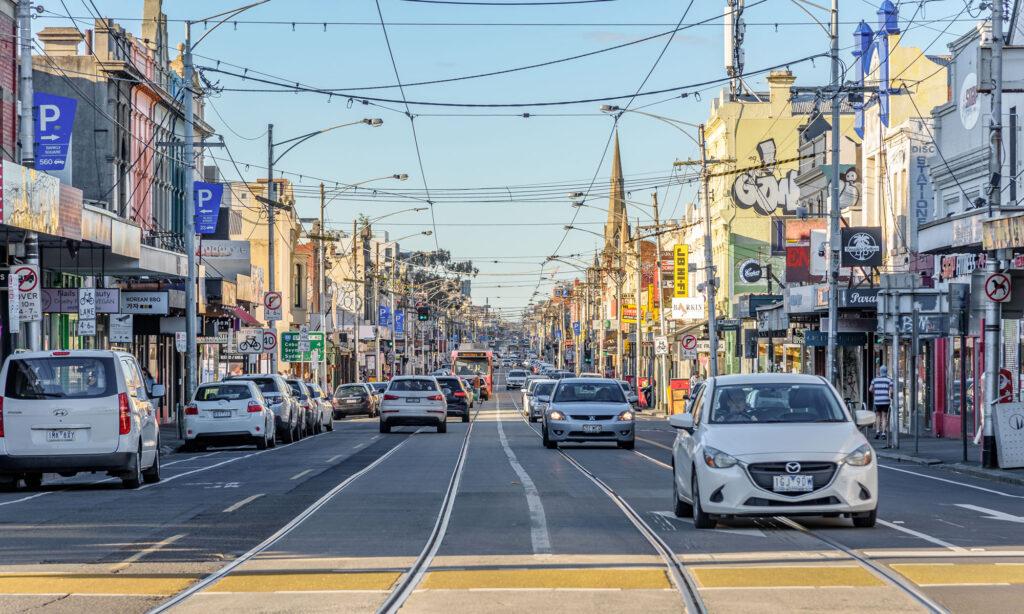 The photo displays a busy intersection on Sydney Rd, Brunswick. The sky is blue and there are lots of shopfronts, with powerlines above.