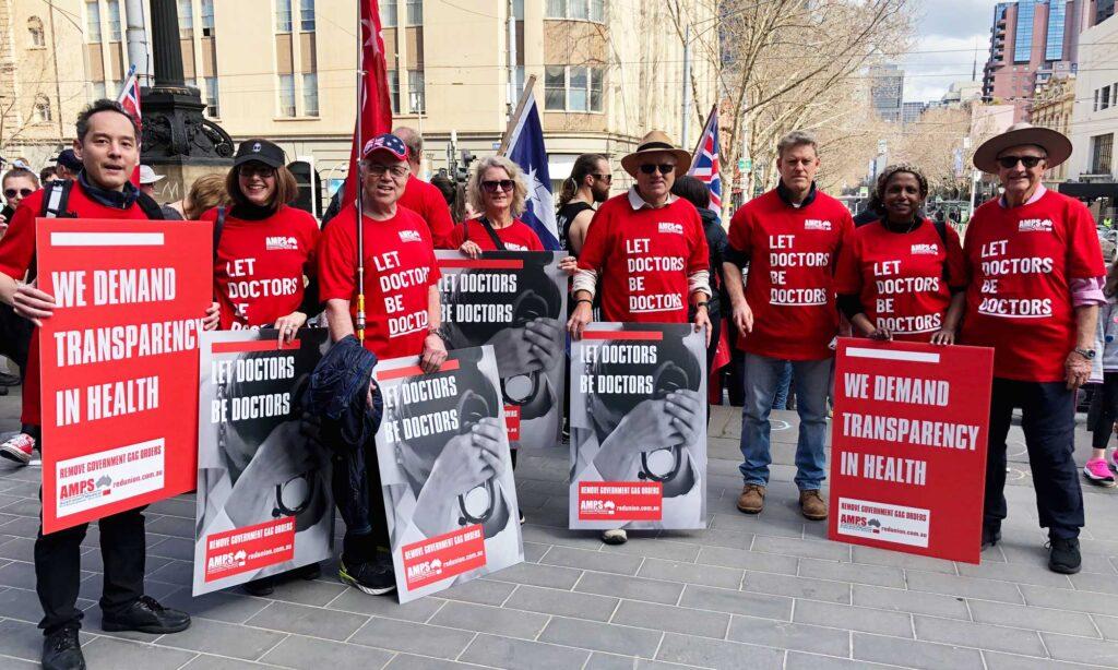 A group of eight protesters representing the AMPS (Australian Medical Professionals Society) dressed in red t-shirts hold protests signs reading 'We demand transparency in health' and 'Let doctors be doctors'. The protesters are standing on the steps of Parliament House, on Spring St in Melbourne's CBD.