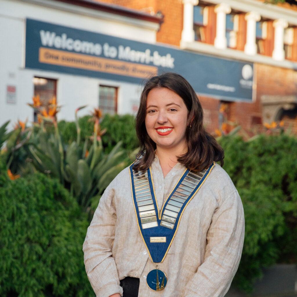 Merri-bek Council mayor, Angelica Panoupolos wears a beige shirt and a blue medal in front of a sign that reads 'Welcome to Merri-bek'
