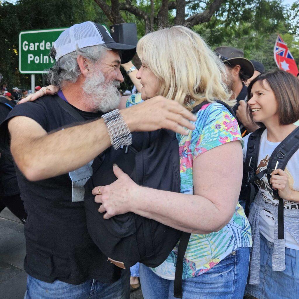 Michael Griffith wears a white and black trucker hat and embraces a woman in a bright Hawaiian style short at a rally.