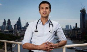 Dr Aseem Malhotra, leaning on a railing. He has a stethoscope around his neck, and wears a light blue shirt. In the background, a city skyline and river.